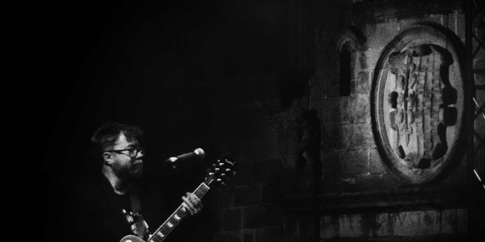 Troy Nahumko at Cáceres Blues Festival Guitarist in the Old Town of Caceres in front of a medieval shield.