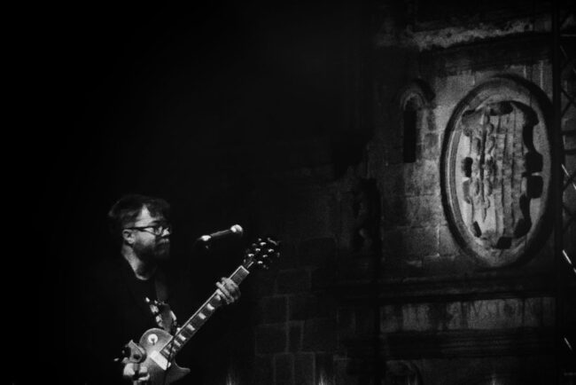 Troy Nahumko at Cáceres Blues Festival Guitarist in the Old Town of Caceres in front of a medieval shield.